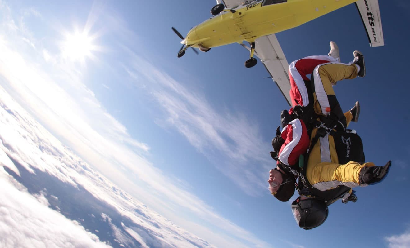 Tandem skydive in freefall after jumping from a yellow airplane above the clouds, with instructor behind the passenger.