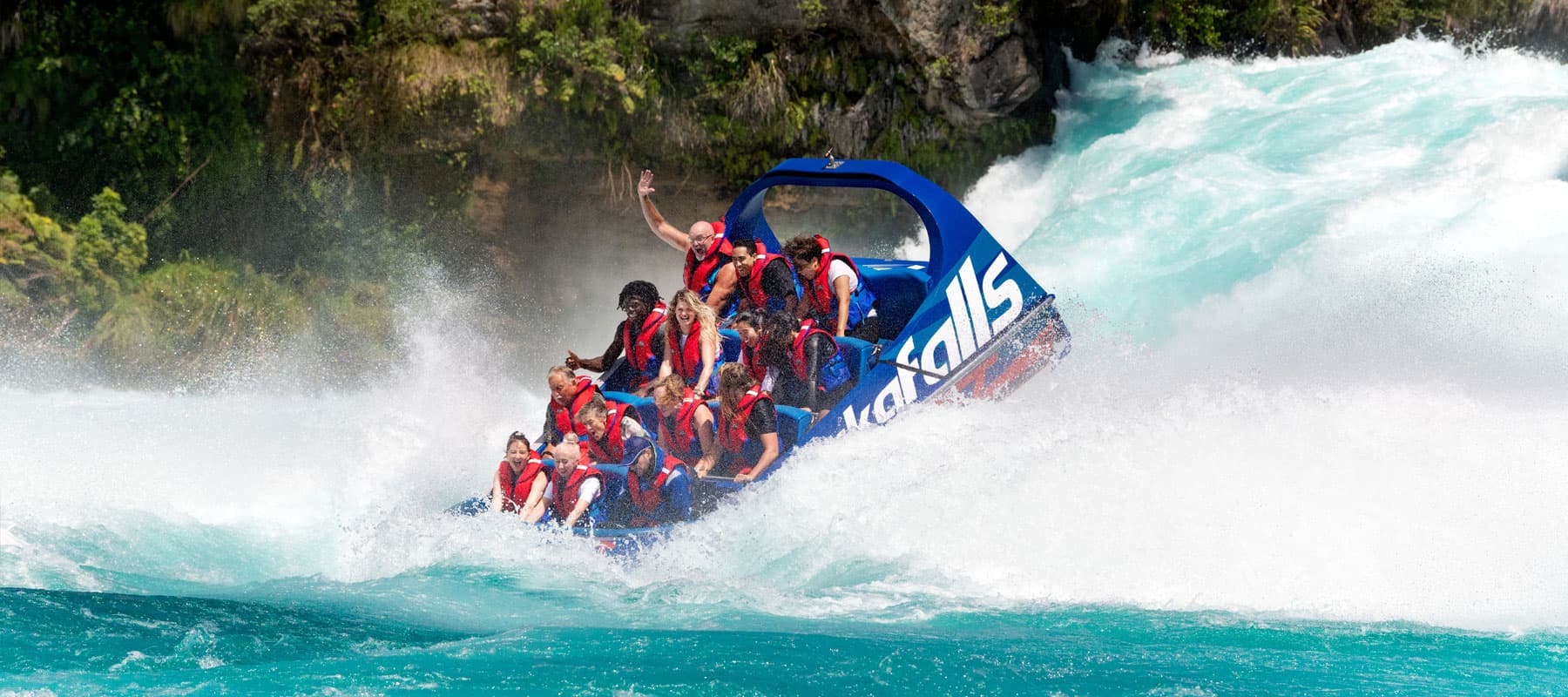  The image shows a blue jet boat labeled "hukafalls" navigating through turbulent whitewater rapids. The passengers, all wearing life jackets, appear to be experiencing the thrill of the ride, with one person raising their hand in excitement. The boat is 