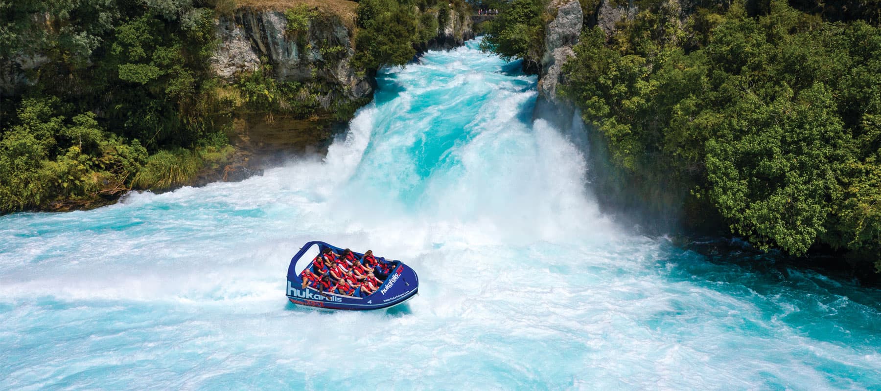 Hukafalls Jet boat filled with passengers at the base of Huka Falls waterfall in Taupo, New Zealand, surrounded by lush green bush and vibrant blue water