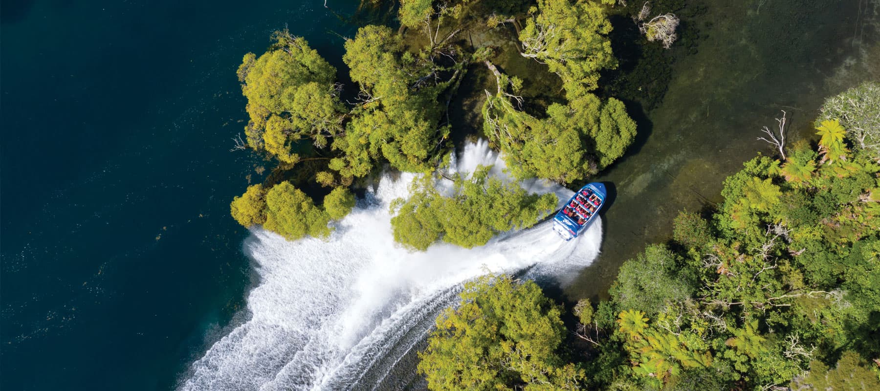  The image shows an aerial view of a blue jet boat labeled "hukafalls JET" making a sharp turn near a cluster of green trees that extend out into the water. The boat leaves a large white wake as it navigates the bend, creating a striking contrast against 