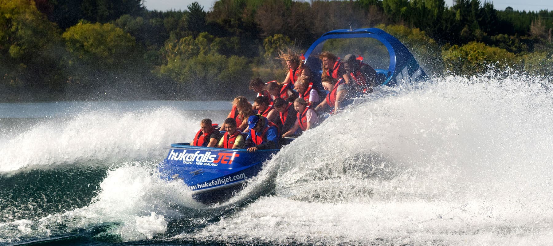  The image shows a blue jet boat labeled "hukafalls JET" filled with passengers wearing red life jackets as they speed through the water. The boat creates a large splash as it moves, with water spraying high into the air. The passengers appear thrilled an