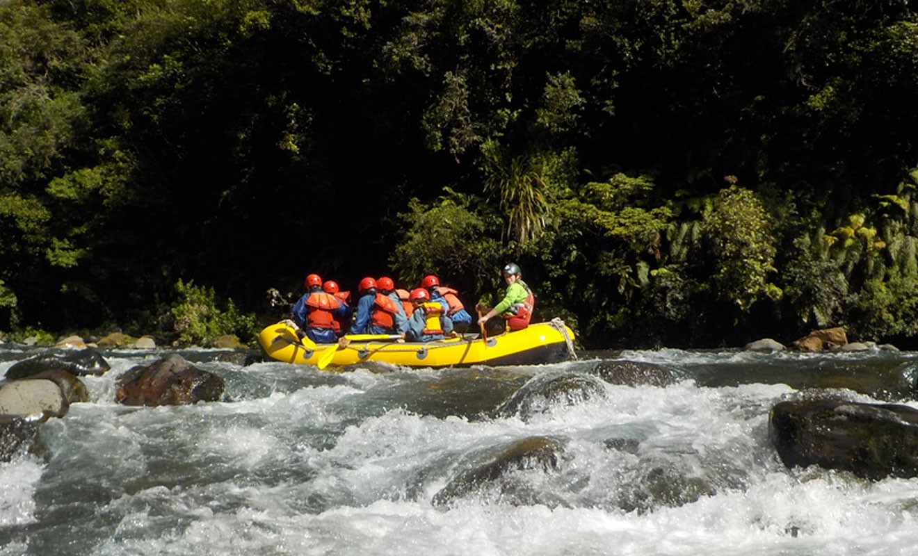  The image shows a group of people rafting down a river in a yellow inflatable raft. The passengers, wearing orange life jackets and helmets, are navigating through rocky rapids, with whitewater surrounding the raft. A guide at the back steers the raft wh
