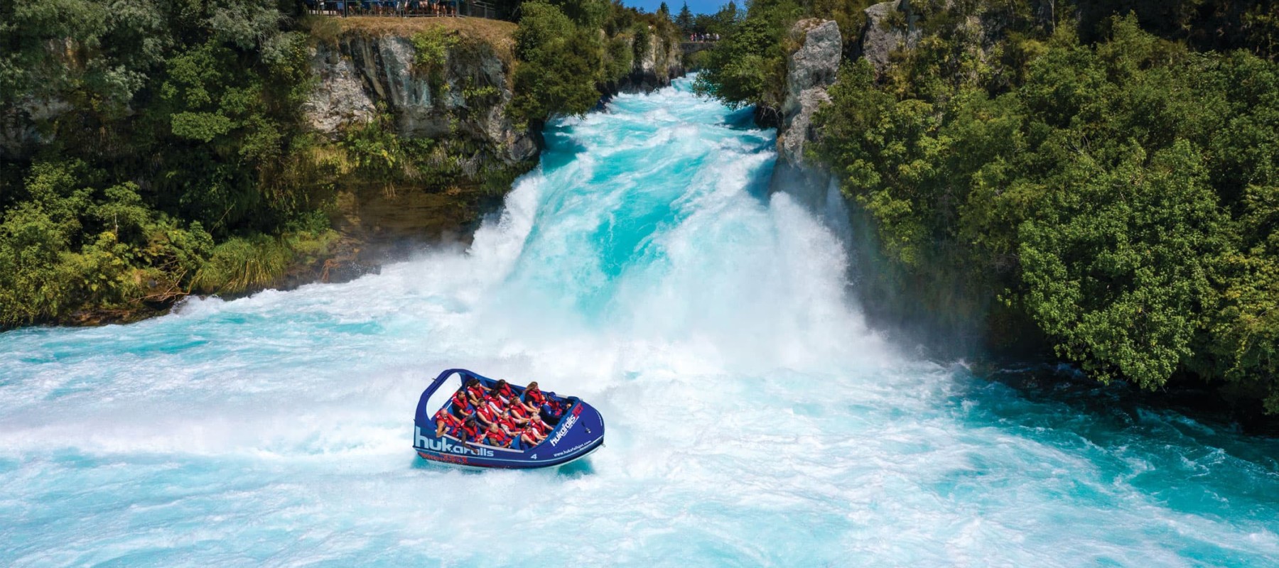 Huka Falls Jet boat at the base of Huka Falls on blue water with green trees along the riverbank in Taupō, New Zealand.