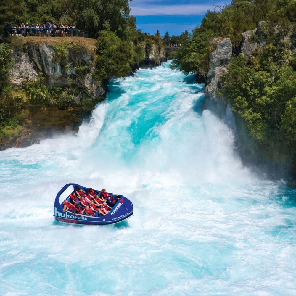 Huka Falls Jet boat at the base of Huka Falls on blue water with green trees along the riverbank in Taupō, New Zealand.