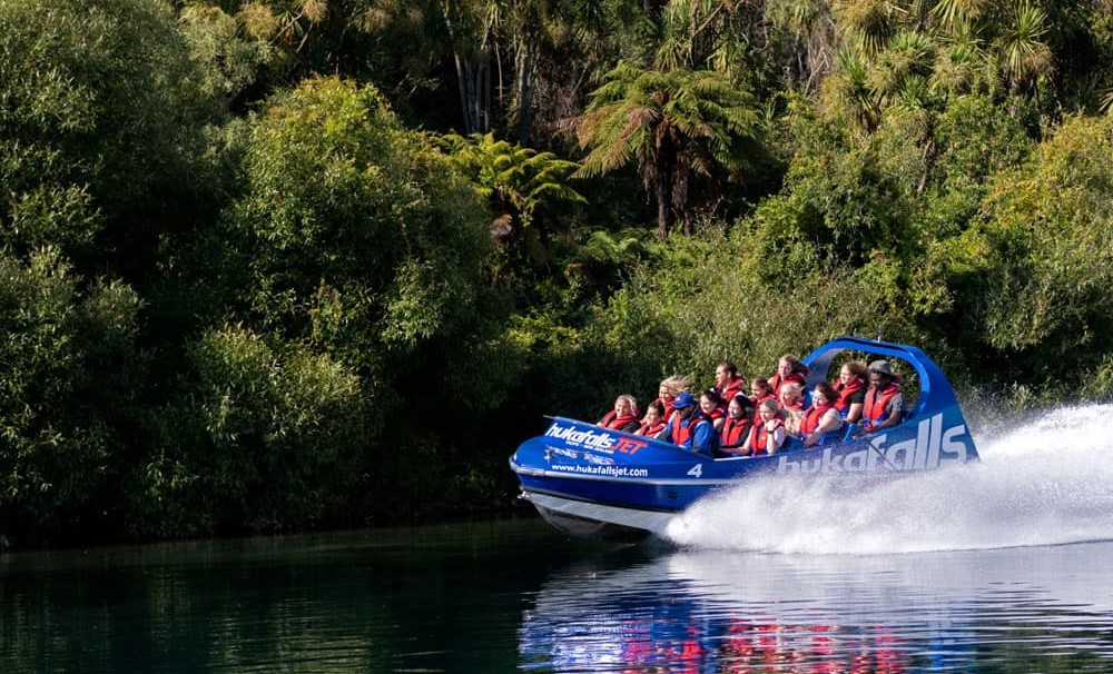 Huka Falls Jet boat with passengers speeding on Waikato River with water spraying behind the boat, surrounded by green trees in Taupō.