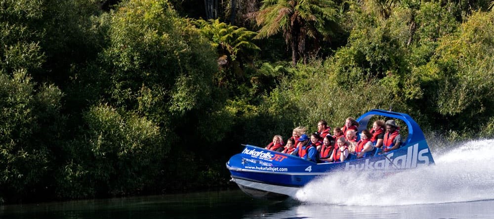 Huka Falls Jet boat with passengers speeding on Waikato River with water spraying behind the boat, surrounded by green trees in Taupō.